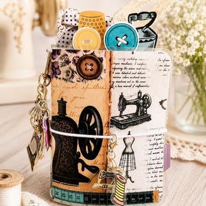May include: A small notebook featuring a vintage sewing theme with a cover displaying a black sewing machine illustration, script text, and a brown button. It includes a metal charm keychain, a white elastic band, and a printed measuring tape border.