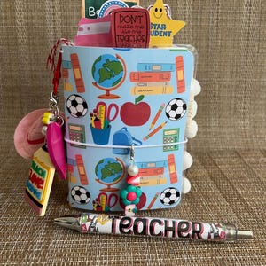 May include: A light blue planner with school-themed illustrations, including globes, books, and soccer balls. A pen with the word "Teacher" is in front of the planner. Charms and decorative accents complete the design.