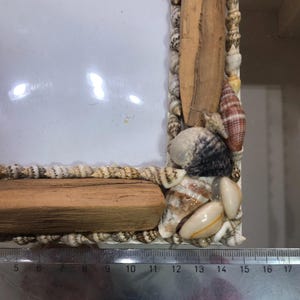 K&ouml;nnte beinhalten: Ein rechteckiger Fotorahmen, verziert mit Muscheln und Treibholzst&uuml;cken. Der Rahmen ist mit Muscheln in verschiedenen Gr&ouml;&szlig;en und Farben verziert, mit einem Abschnitt aus hellem Holz oben und unten. Der Rahmen ist etwa 15 cm breit.