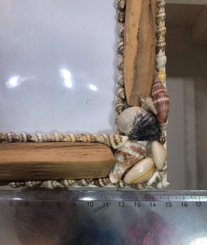 K&ouml;nnte beinhalten: Ein rechteckiger Fotorahmen, verziert mit Muscheln und Treibholzst&uuml;cken. Der Rahmen ist mit Muscheln in verschiedenen Gr&ouml;&szlig;en und Farben verziert, mit einem Abschnitt aus hellem Holz oben und unten. Der Rahmen ist etwa 15 cm breit.