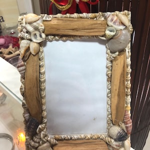 Può includere: Una cornice rettangolare decorata con conchiglie e legno. La cornice ha un bordo in legno marrone chiaro ed è adornata con una varietà di conchiglie di diverse forme, dimensioni e colori. È un oggetto decorativo.