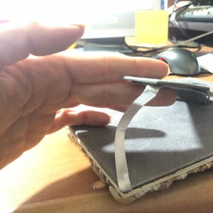 K&ouml;nnte beinhalten: Eine graue, rechteckige Fliese mit einem wei&szlig;en Klebestreifen. Die Fliese hat eine strukturierte Oberfl&auml;che und wird von einer Hand gehalten. Die Kanten der Fliese sind mit einem hellfarbigen Material versehen.