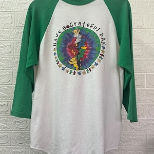 May include: White and green raglan shirt with the text "Have a Grateful Day" and a graphic of dancing bears. The shirt has green sleeves and a white body. The graphic features a tie-dye background.