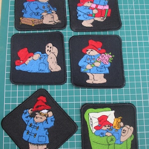 May include: Six embroidered fabric patches featuring a bear character wearing a red hat and blue coat. The designs show the bear sitting on a suitcase, holding gifts, lying down, holding flowers, adjusting a hat, and sitting in a green armchair.