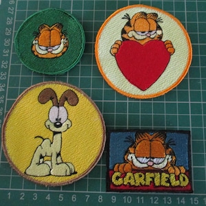 May include: Four embroidered patches featuring Garfield characters. One small circular green patch with Garfield's face, one large circular patch with an orange border showing Garfield holding a red heart, one large circular yellow patch with Odie, and one rectangular blue patch with Garfield and the text GARFIELD.