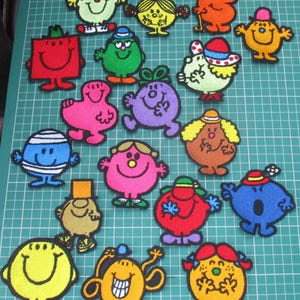 May include: A selection of colourful embroidered patches depicting cartoon characters with smiling faces. The patches are in various shapes and colours, including red, blue, yellow, and pink, arranged on a green cutting mat.