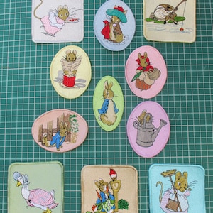 May include: A collection of ten embroidered fabric patches featuring various animal characters in clothing, including rabbits, mice, and a goose. The patches are square or oval in shape, displaying colourful illustrations of animals engaged in activities.