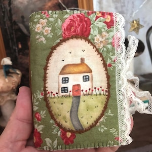 Op de afbeelding: Een klein notitieboekje bekleed met stof met een groene bloemenprint en rode rozen. Het midden toont een ovaal embleem met een handgeschilderd huisje, een rode deur en een pad. De rand is versierd met witte kanten afwerking en een lint.