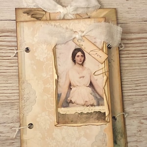 May include: A vintage-style scrapbook with a white lace bow and a picture of an angel with the word "Love" on a tag. The scrapbook has a distressed look with a floral pattern.