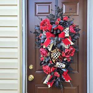 May include: A decorative door wreath featuring red roses, black leaves, and a large red bow. Accents include black and white checkered ribbon and a decorative pumpkin. The wreath is displayed on a brown door.