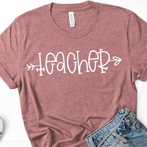 May include: A dusty rose-colored t-shirt with the word "TEACHER" in white script, with an arrow design. The shirt is made of a soft material and is paired with blue denim jeans.