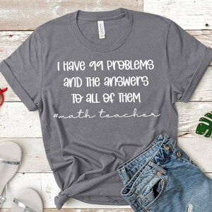 Op de afbeelding: Grijs T-shirt met witte tekst die luidt "Ik heb 99 problemen en de antwoorden op alles #wiskundeleraar".