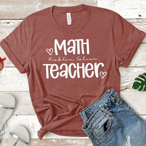 Op de afbeelding: Een maronkleurig T-shirt met witte tekst die "Math Problem Solver Teacher" luidt met een hartje aan weerszijden van de tekst.