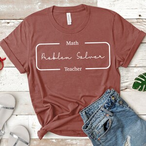 Op de afbeelding: Een maronkleurig T-shirt met witte tekst die "Math Problem Solver Teacher" in een rechthoekige doos zegt.