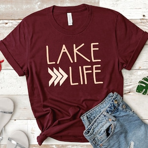 May include: A maroon t-shirt with the text "LAKE LIFE" in a peach color. The text is stylized with three arrows pointing to the right.