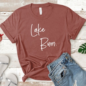 May include: A rust-colored t-shirt with the text "Lake Bum." printed in white. The shirt is laid out on a wooden surface with a pair of sandals and a pair of jeans.