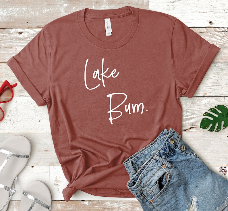 May include: A rust-colored t-shirt with the text "Lake Bum." printed in white. The shirt is laid out on a wooden surface with a pair of sandals and a pair of jeans.