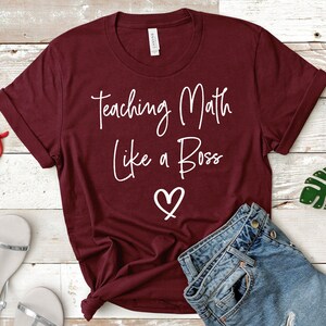 Op de afbeelding: Een maroon t-shirt met de tekst "Teaching Math Like a Boss" in witte letters en een wit hart.