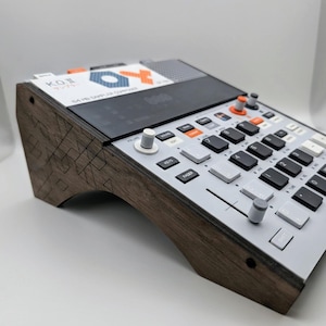 May include: A music sampler device with a light grey control panel featuring black and white buttons, orange accents, and a digital display labelled KO II. The unit is mounted on a dark brown wooden stand with a geometric laser-etched pattern.
