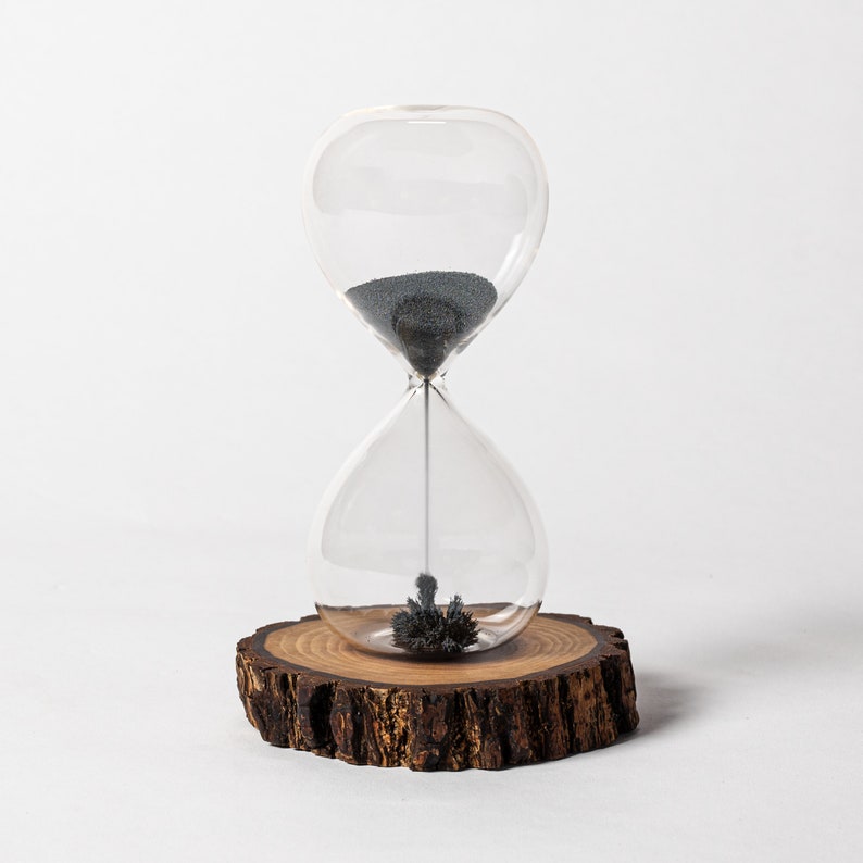 May include: A glass hourglass with black sand sits on a wooden base. The sand is slowly flowing through the hourglass, creating a mesmerizing effect.
