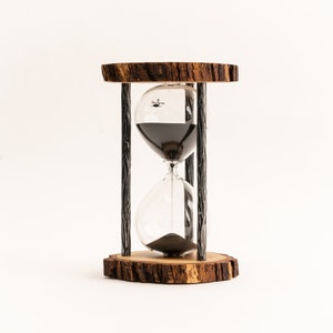 Könnte beinhalten: Eine Sanduhr aus Holz mit schwarzem Sand, einer Glasbirne und einem Metallrahmen. Die Sanduhr steht auf einem Holzsockel.