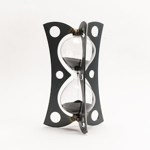 Può includere: Una clessidra in metallo nero con un bulbo in vetro trasparente e sabbia nera. La clessidra è su uno sfondo bianco.