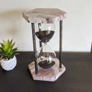 May include: A marble hourglass with a black sand timer inside. The hourglass is on a marble base with a hexagonal shape. The base has a brown and white marbled pattern.