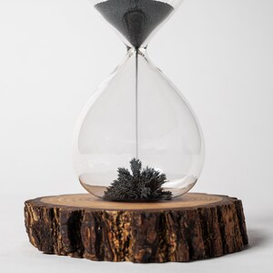 May include: A glass hourglass with black sand sits on a wooden base. The sand is clumped together in the bottom of the hourglass.