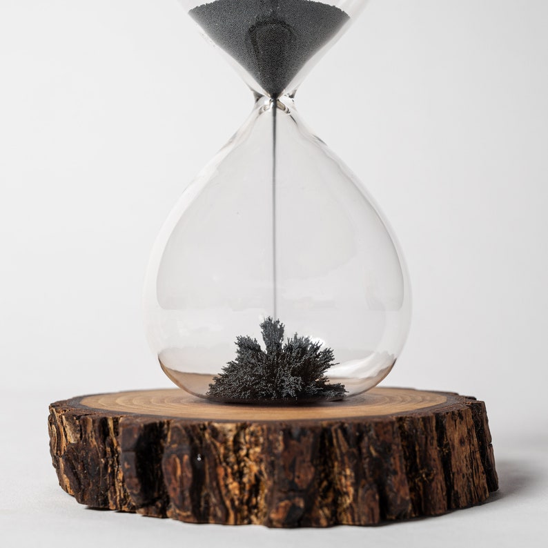 May include: A glass hourglass with black sand sits on a wooden base. The sand is clumped together in the bottom of the hourglass.