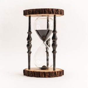 Könnte beinhalten: Eine schwarze Sanduhr mit einem Holzsockel. Die Sanduhr ist mit schwarzem Sand gefüllt und hat eine klare Glasbirne.