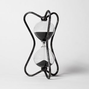 Puede incluir: Un reloj de arena negro con un marco de metal retorcido. El reloj de arena está lleno de arena negra y está sobre una superficie blanca.
