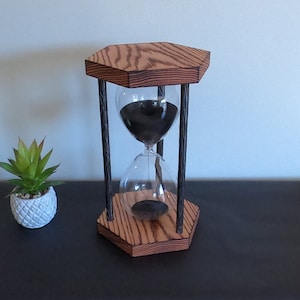 May include: A wooden hourglass with black sand and a hexagonal base. The hourglass is made of dark wood with a wood grain pattern. The hourglass is sitting on a black surface.