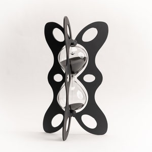 Puede incluir: Reloj de arena negro con una bombilla de vidrio transparente y arena negra. El reloj de arena se mantiene en su lugar mediante un marco calado negro y decorativo. El marco tiene un diseño curvo único con recortes ovalados. Es un artículo decorativo.