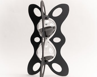 Op de afbeelding: Zwarte zandloper met een heldere glazen bol en zwart zand. De zandloper wordt op zijn plaats gehouden door een zwart, decoratief, opengewerkt frame. Het frame heeft een uniek, gebogen ontwerp met ovale uitsparingen. Het is een decoratief item.