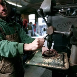 May include: A person operating a vintage drill press with a wood block clamped in place. The drill press is labeled "Canadian".