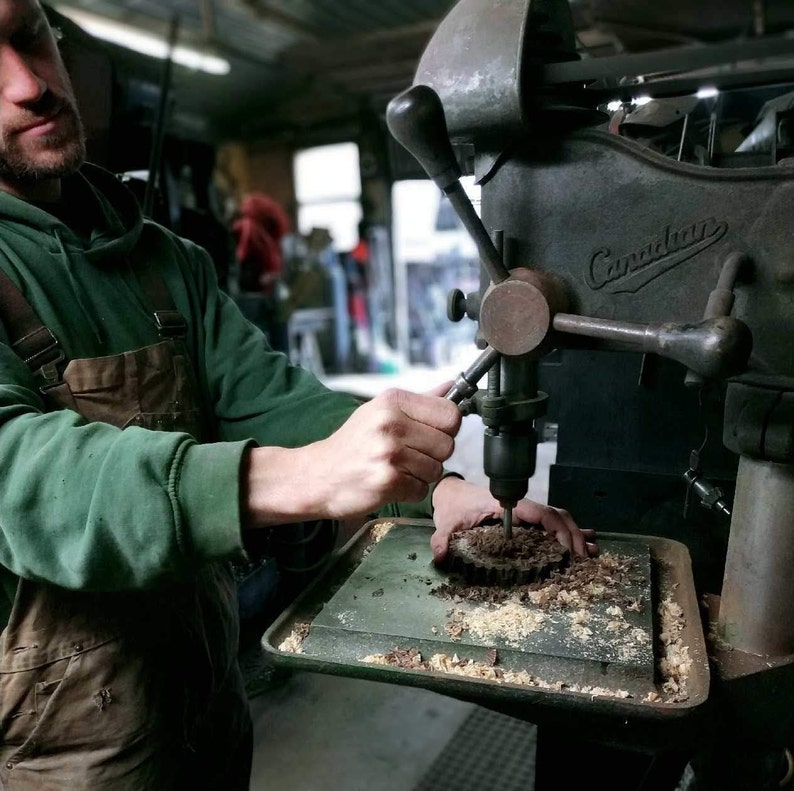 May include: A person operating a vintage drill press with a wood block clamped in place. The drill press is labeled "Canadian".