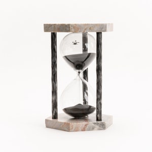 May include: A black sand hourglass with a marble base and metal frame. The hourglass is sitting on a white surface.