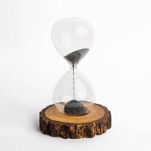 May include: A glass hourglass with black sand sits on a wooden base. The sand is flowing from the top bulb to the bottom bulb.