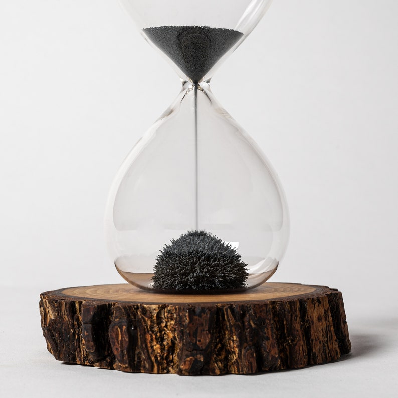 May include: A glass hourglass with black sand sits on a wooden base. The sand is clumped together in the bottom of the hourglass.