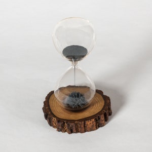May include: A glass hourglass with black sand on a wooden base. The hourglass is sitting on a wooden base that looks like a tree stump.