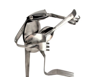 May include: A silver frog sculpture made from repurposed silverware. The frog is playing a guitar made from a spoon and fork.