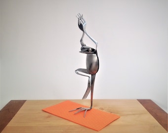 May include: A whimsical sculpture of a frog crafted from silverware, posed in a yoga stance. The frog is made from spoons and forks, with a fork forming the head and arms. It stands on an orange mat, set against a light background.