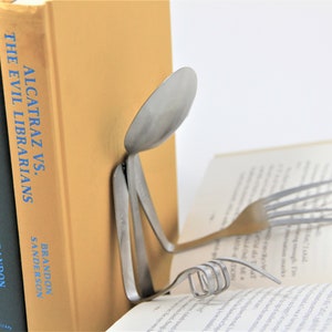 Puede incluir: Una cuchara y un tenedor de plata est&aacute;n doblados en forma de persona leyendo un libro. El libro se titula "Alcatraz vs. The Evil Librarians" de Brandon Sanderson. El libro est&aacute; apoyado por otros dos libros, uno titulado "The Scrivener's Bones" de Brandon Sanderson.