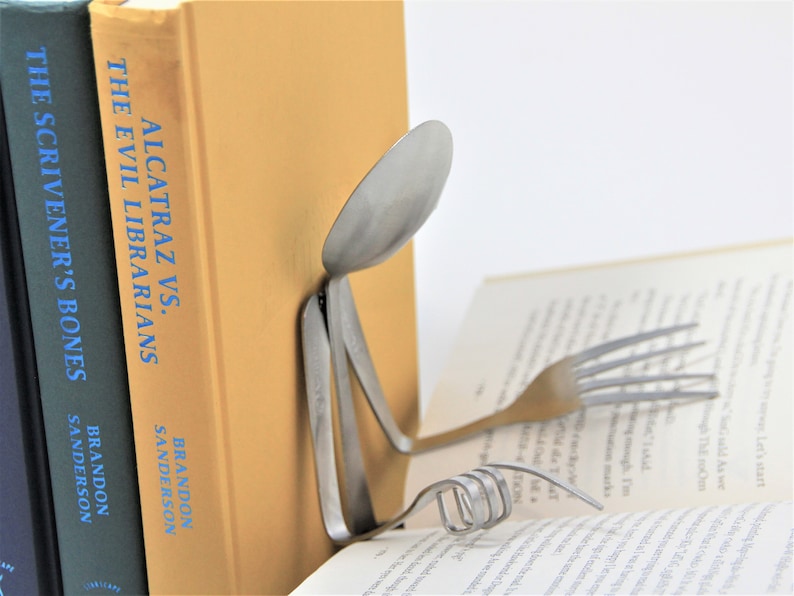 Puede incluir: Una cuchara y un tenedor de plata est&aacute;n doblados en forma de persona leyendo un libro. El libro se titula "Alcatraz vs. The Evil Librarians" de Brandon Sanderson. El libro est&aacute; apoyado por otros dos libros, uno titulado "The Scrivener's Bones" de Brandon Sanderson.