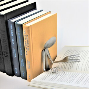 Puede incluir: Un sujetalibros hecho de una cuchara y un tenedor, sosteniendo un libro. El sujetalibros est&aacute; hecho de metal y tiene la forma de una persona leyendo. El libro est&aacute; abierto y tiene un fondo blanco. El sujetalibros est&aacute; sentado sobre una pila de libros con los t&iacute;tulos "Alcatraz vs. The Evil Librarians", "The Scrivener's Bones", y "The Dark Talent" de Brandon Sanderson.