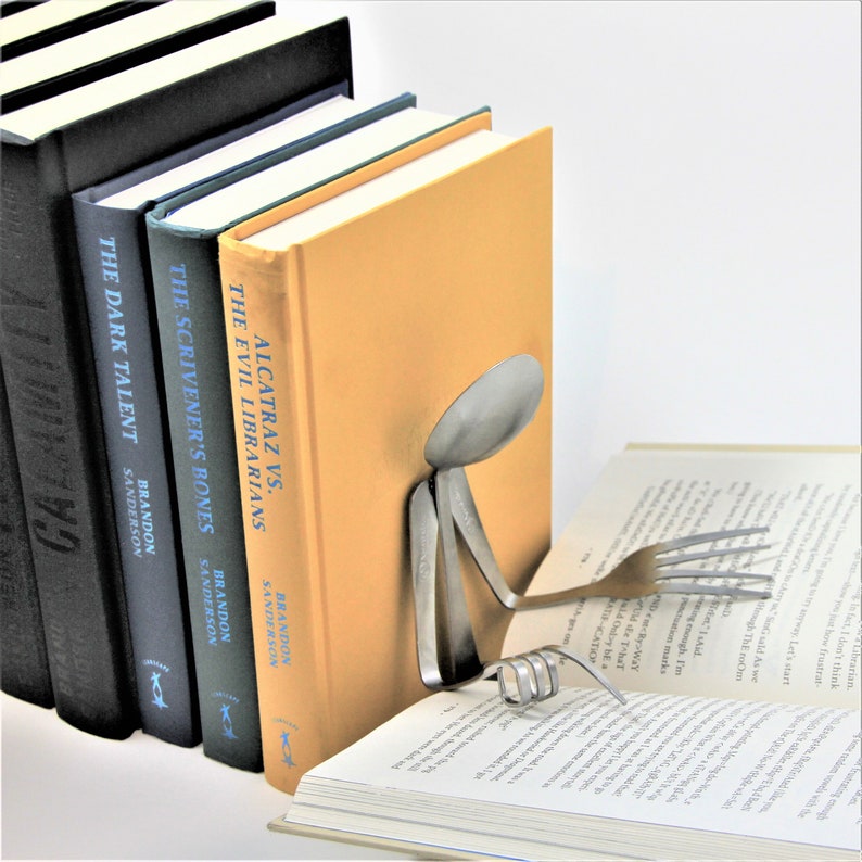 Puede incluir: Un sujetalibros hecho de una cuchara y un tenedor, sosteniendo un libro. El sujetalibros est&aacute; hecho de metal y tiene la forma de una persona leyendo. El libro est&aacute; abierto y tiene un fondo blanco. El sujetalibros est&aacute; sentado sobre una pila de libros con los t&iacute;tulos "Alcatraz vs. The Evil Librarians", "The Scrivener's Bones", y "The Dark Talent" de Brandon Sanderson.