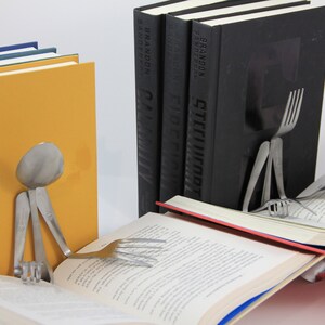 Puede incluir: Dos sujetalibros hechos de cubiertos, con forma de personas, sosteniendo libros. Un sujetalibros est&aacute; hecho de una cuchara y tenedores, el otro de un tenedor y un cuchillo. Los sujetalibros sostienen dos libros abiertos.