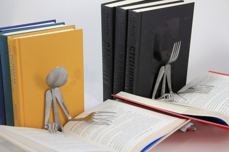 Puede incluir: Dos sujetalibros hechos de cubiertos, con forma de personas, sosteniendo libros. Un sujetalibros est&aacute; hecho de una cuchara y tenedores, el otro de un tenedor y un cuchillo. Los sujetalibros sostienen dos libros abiertos.