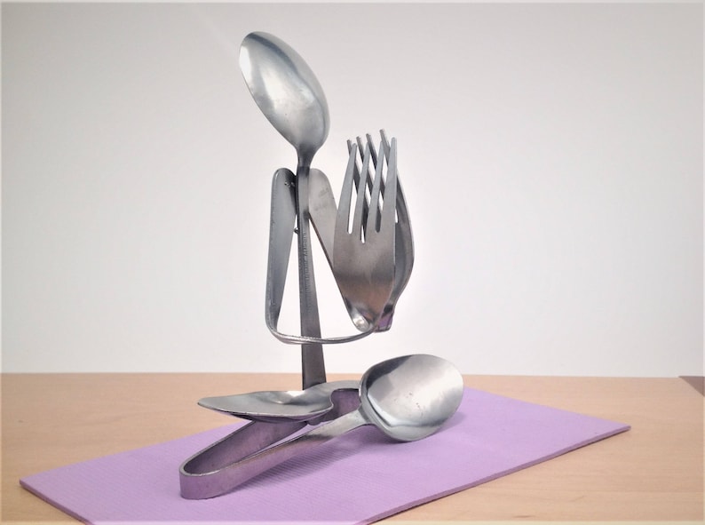 May include: A sculpture made from silverware, including a spoon, fork, and tongs. The sculpture is shaped like a person with their hands raised in prayer. The sculpture is sitting on a purple surface.