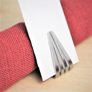 May include: Silver napkin ring with a modern design. The ring is made of multiple thin strips of metal that are layered and curved to create a unique shape.